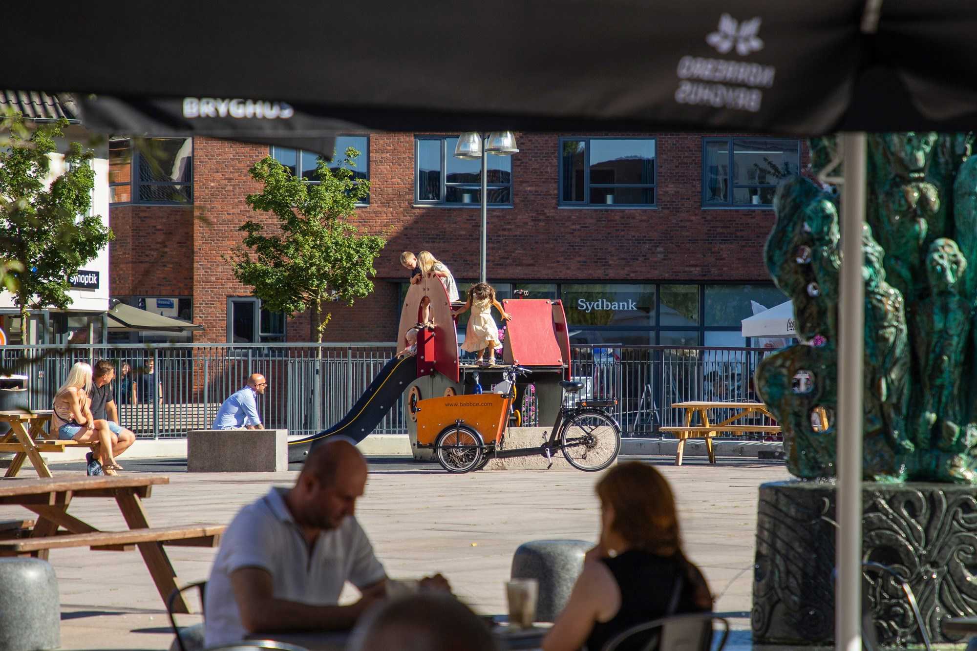 Par sidder på café foran børn der leger på legeplads Ved Sydbank i Næstved Bymidte