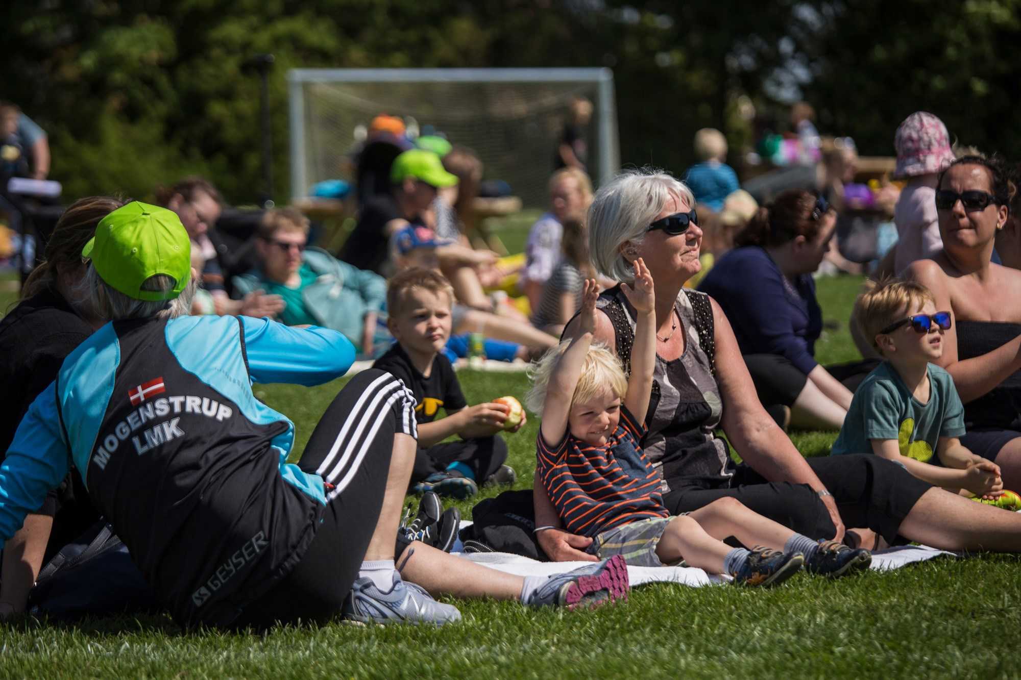 Børn og voksne sidder på græsset og ser på Fodboldkamp i Mogenstrup