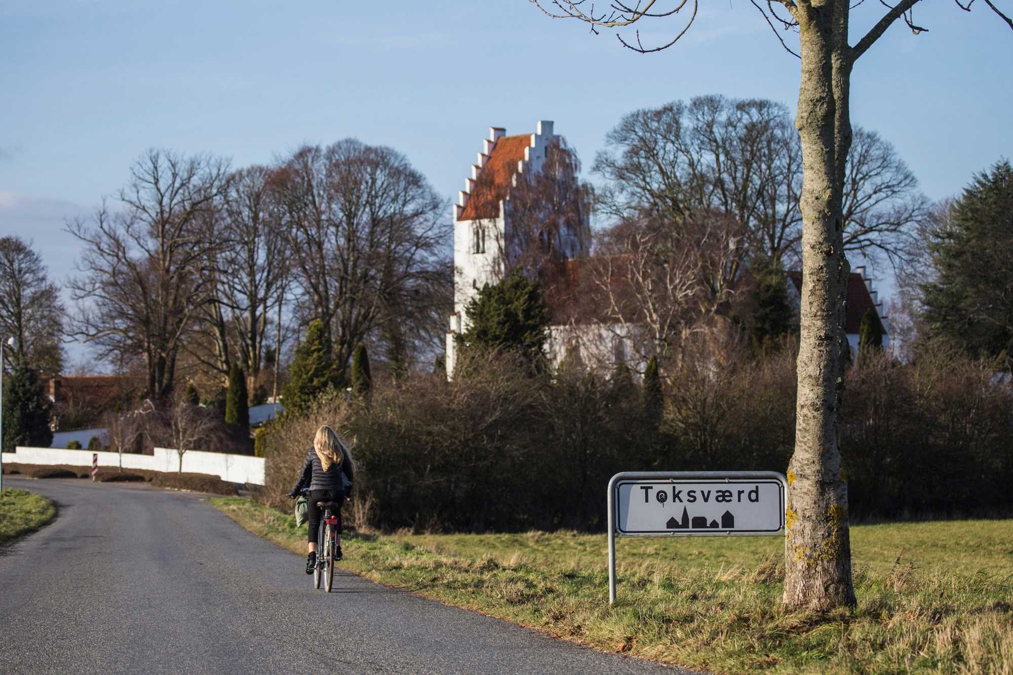 En pige er cyklet forbi byskiltet på vej ind til Toksværd med kirken på hendes højre side