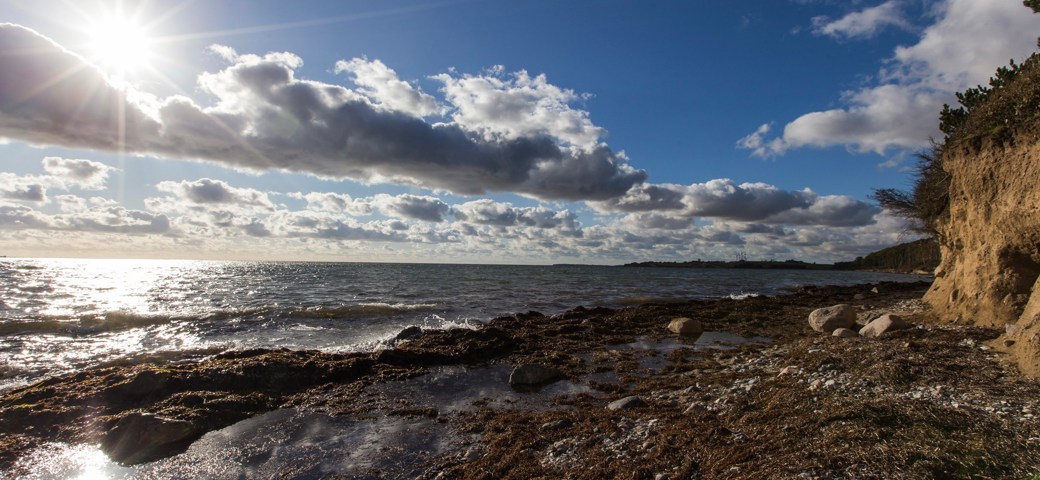 Solen skinner igennem et let skydække imens bølgerne slår ind på kysten ved Karrebæksminde