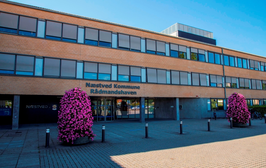 På billedet er der en lang 3 etagers gul murstensbygning, med lange vandrette glaspartier. Foran er der flisebelagt og der er placeret to lilla buske i krukker. På bygningen står der Næstved Kommune Rådmandshaven.
